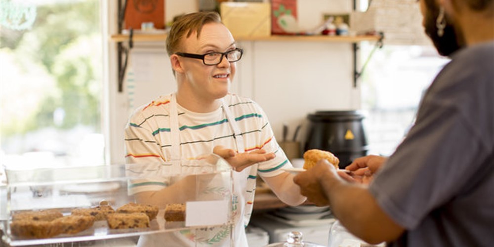 Young man with Downs syndrome working in a cafe serving a customer from behind a display counter