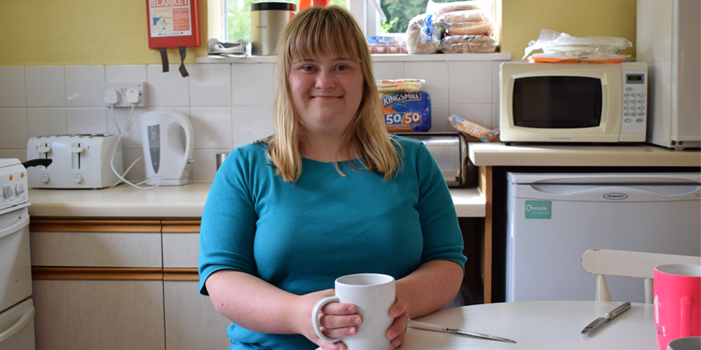 Young woman with Down Syndrome sitting at a kitchen table holding a mug, smiling at the camera