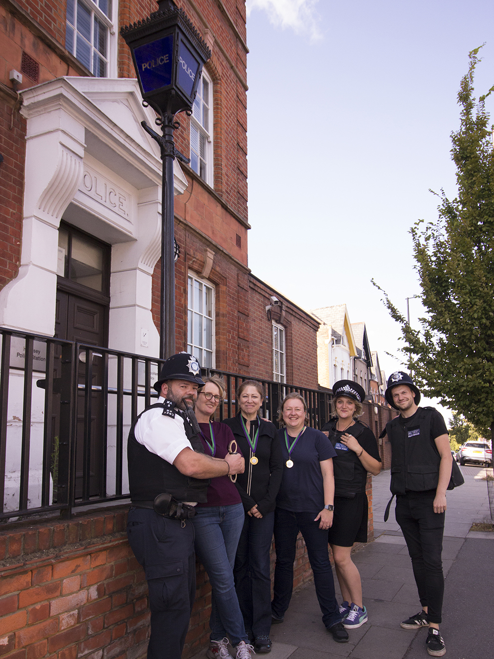 6 people are stood outside a police station, three in police costumes and three wearing medals for completing a challenge